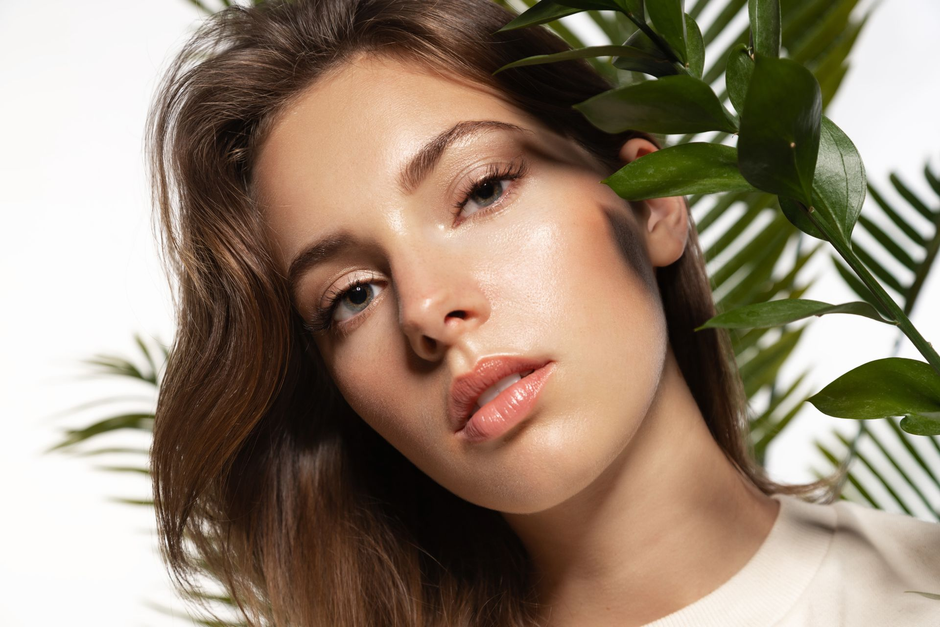 Close-up portrait of a person with brown hair beside green leaves, softly lit against a white background