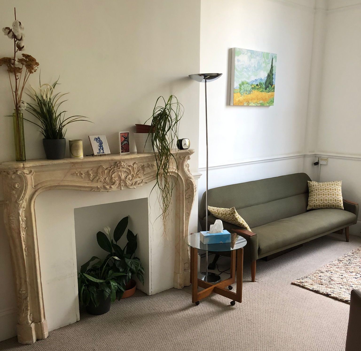 A cozy room with a fireplace, sofa, art, and plants. Light-colored walls and carpet.