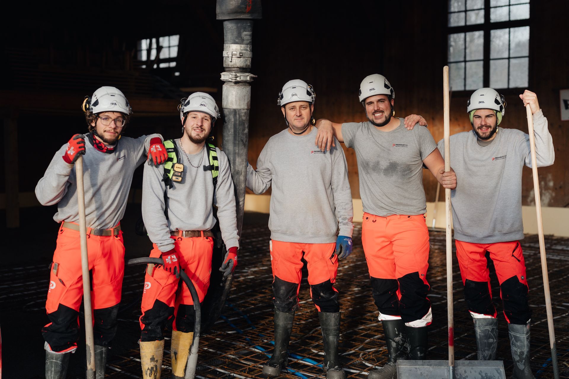 Cinq ouvriers casqués et vêtus de pantalons orange posent avec des pelles à l'intérieur d'un bâtiment industriel sombre.