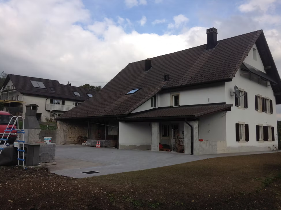 Maison blanche au toit marron et abri voiture dans une allée rurale sous un ciel nuageux