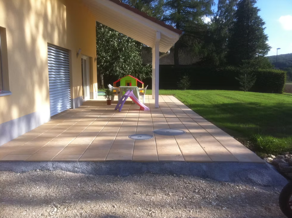 Patio d'une maison jaune avec un petit toboggan coloré pour enfants sous un porche couvert, à côté d'une pelouse.