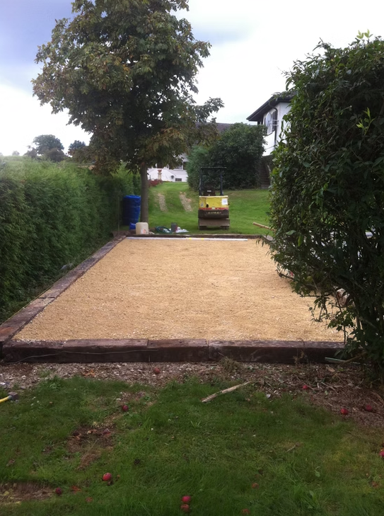 Terrasse rectangulaire en gravier beige bordée de briques, entre de hautes haies, avec un arbre et une pelouse au-delà.