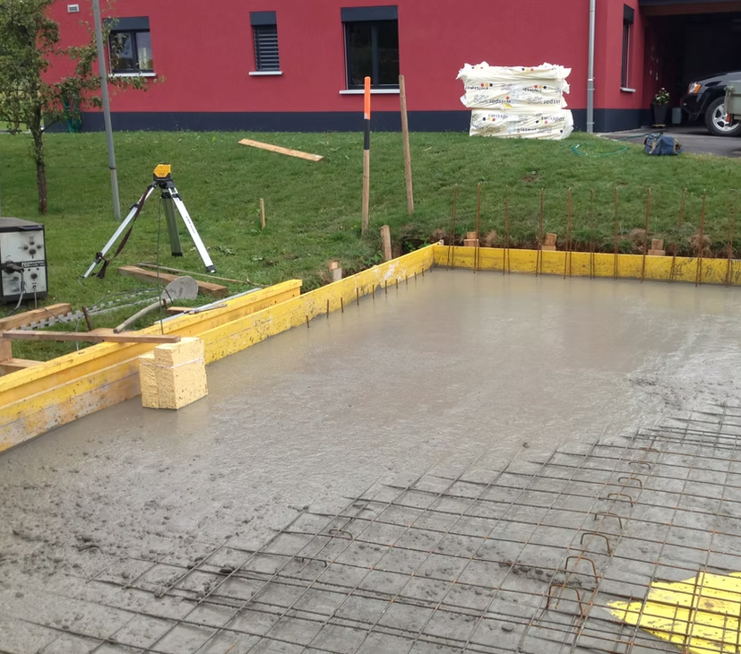Fondation en dalle de béton en construction, avec armatures, coffrage en bois et matériel topographique à proximité.