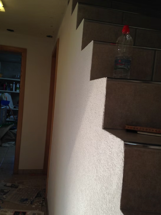 Escalier intérieur faiblement éclairé, avec une bouteille d'eau à bouchon rouge sur la rampe et une porte en arrière-plan.