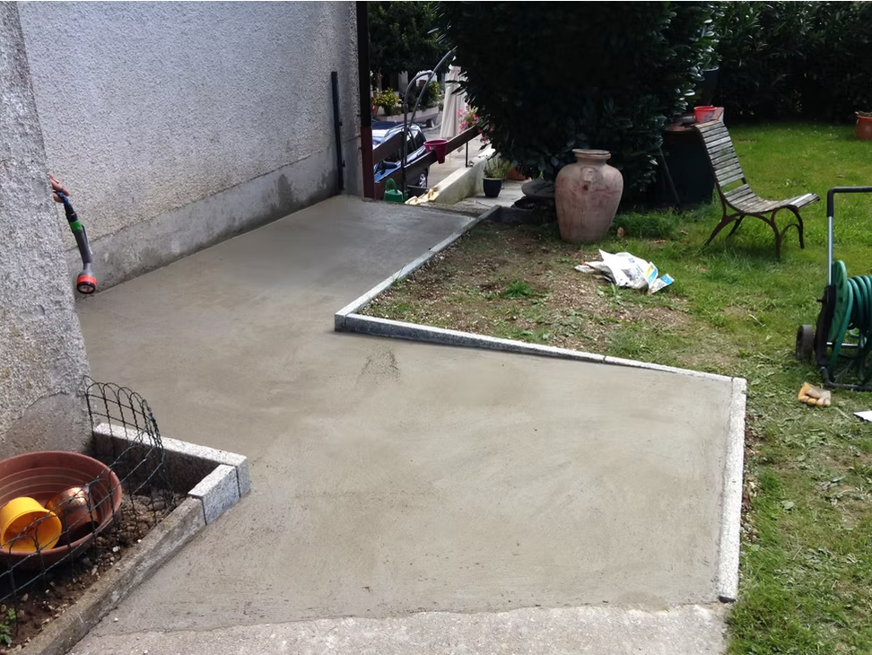 Terrasse en béton jouxtant une pelouse avec un pot en terre cuite, une chaise, un enrouleur de tuyau d'arrosage