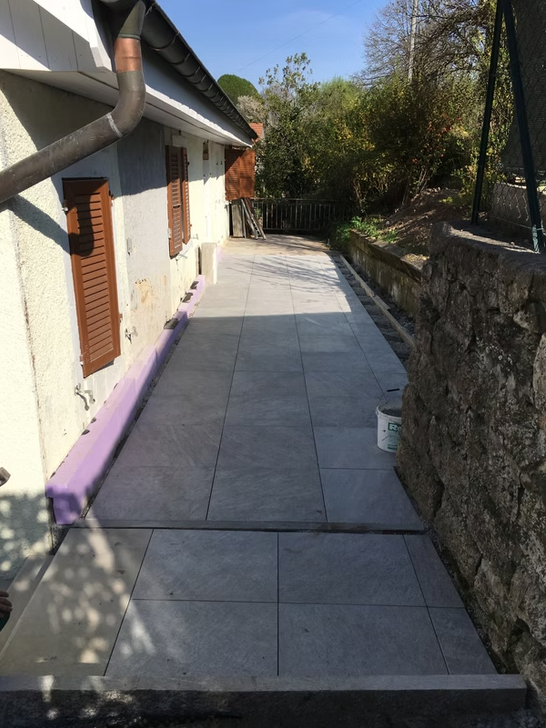 Étroite allée extérieure longeant une maison, avec un pavage carrelé et un mur de pierre sur la droite.