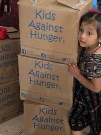 A little Guatemalan girl is holding a cardboard box that says kids against hunger.