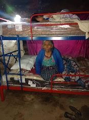 An elderly woman is sitting on top of a bunk bed.