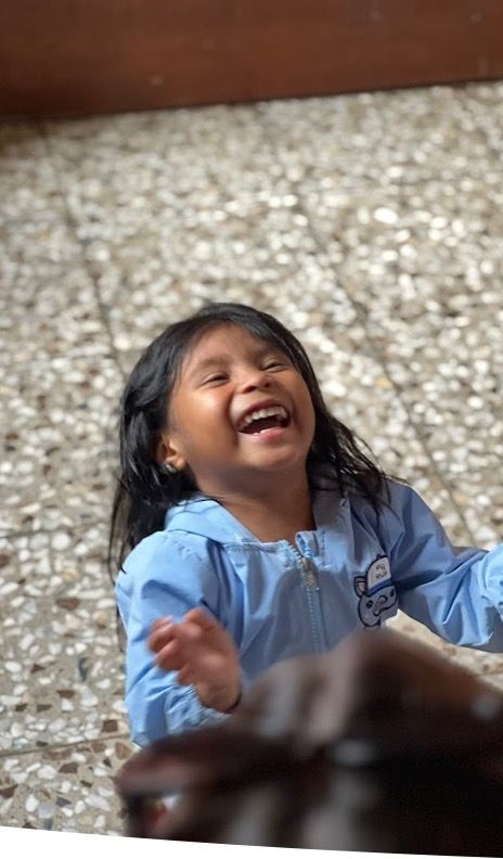 A little GUATELAMALN girl in a blue jacket is laughing while sitting on the floor.