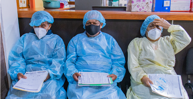 Three nurses wearing masks and gowns are sitting in a waiting room.