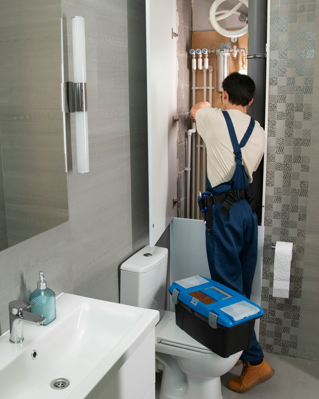 Person in blue overalls reaching into a bathroom cabinet above a toilet and sink.