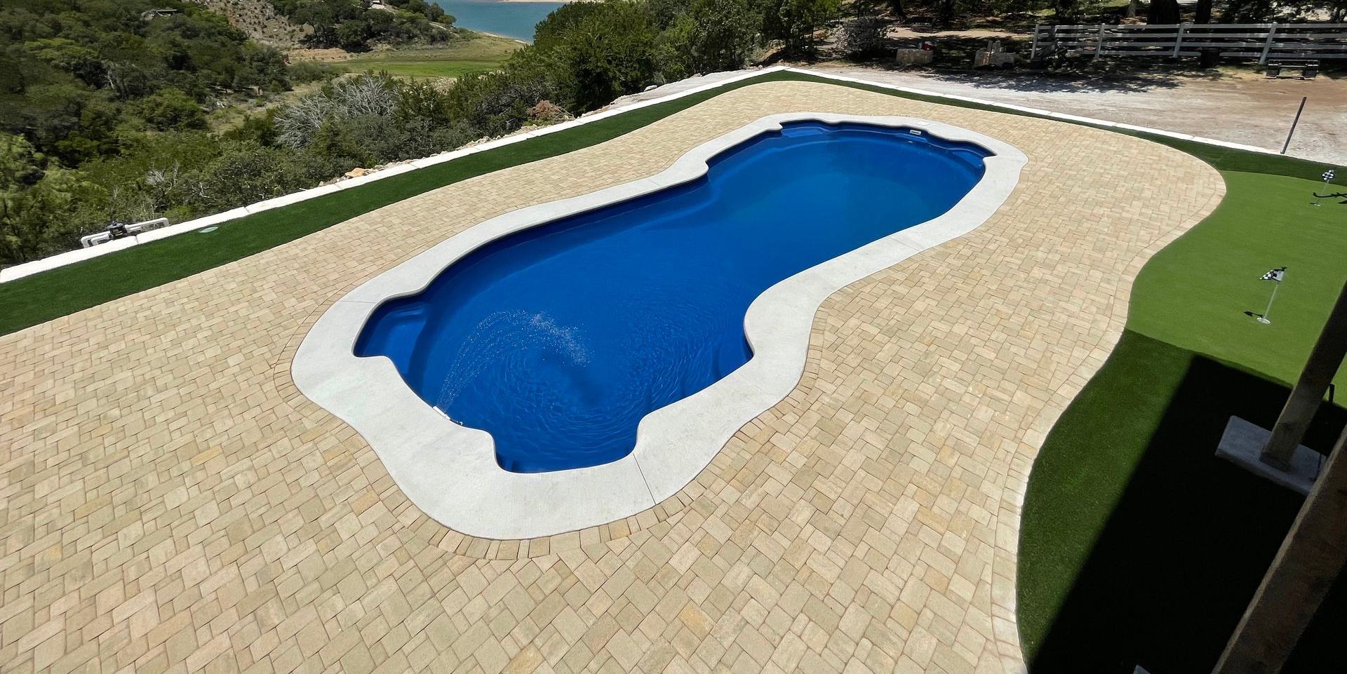 A bright blue swimming pool with curved steps, bordered by a light-colored stone deck and a lush green lawn.