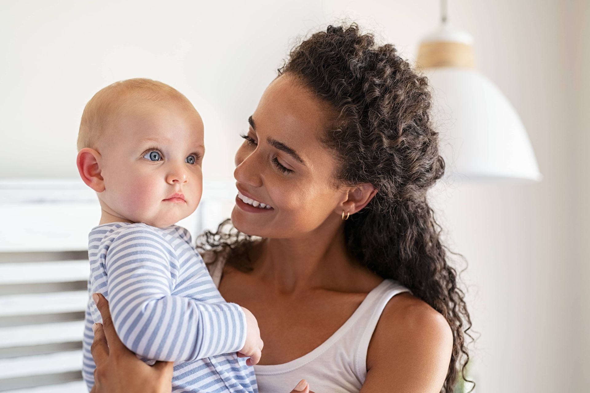 Woman smiling, holding baby in a light-filled room. Baby in blue striped shirt looks toward woman.