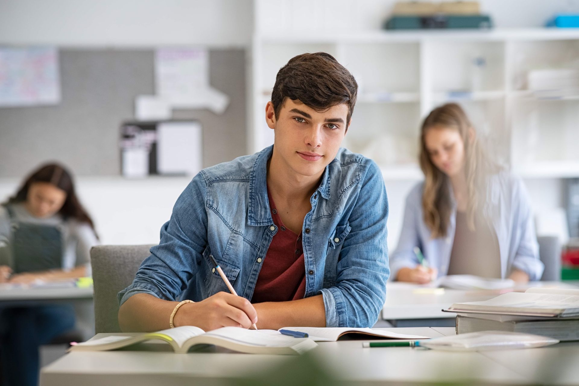 Young person writing at a desk, looking at the camera in a classroom, other students in background.