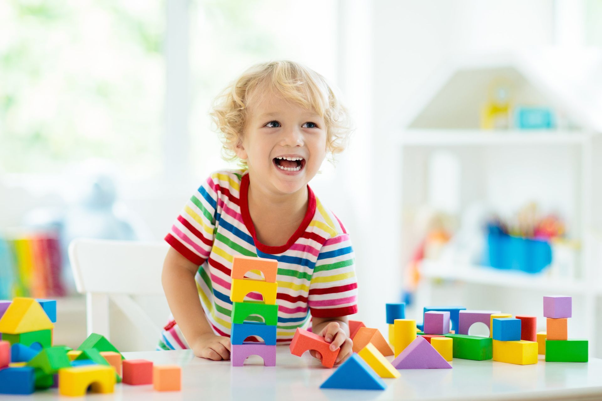 Boy building with colorful blocks on a white table; smiling and laughing in a bright room.