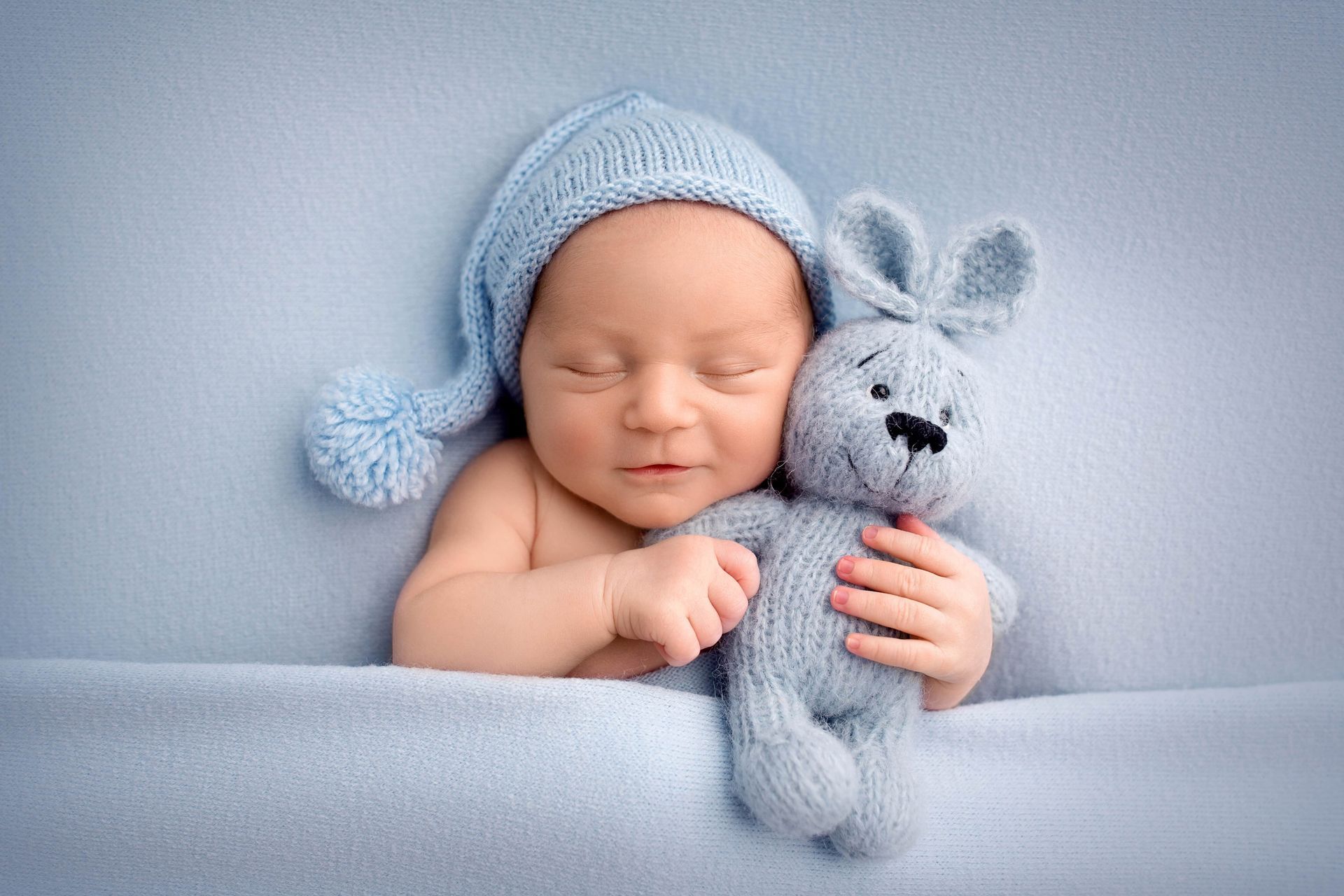 Baby in a blue knit hat, lying on blue blanket, clutching a blue stuffed bunny.