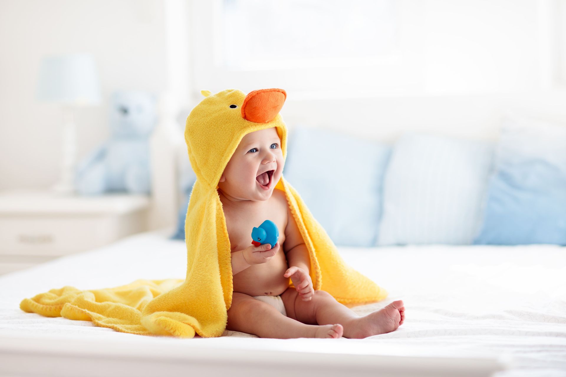 Smiling baby in yellow duck hooded towel sitting on a bed, holding blue toy.
