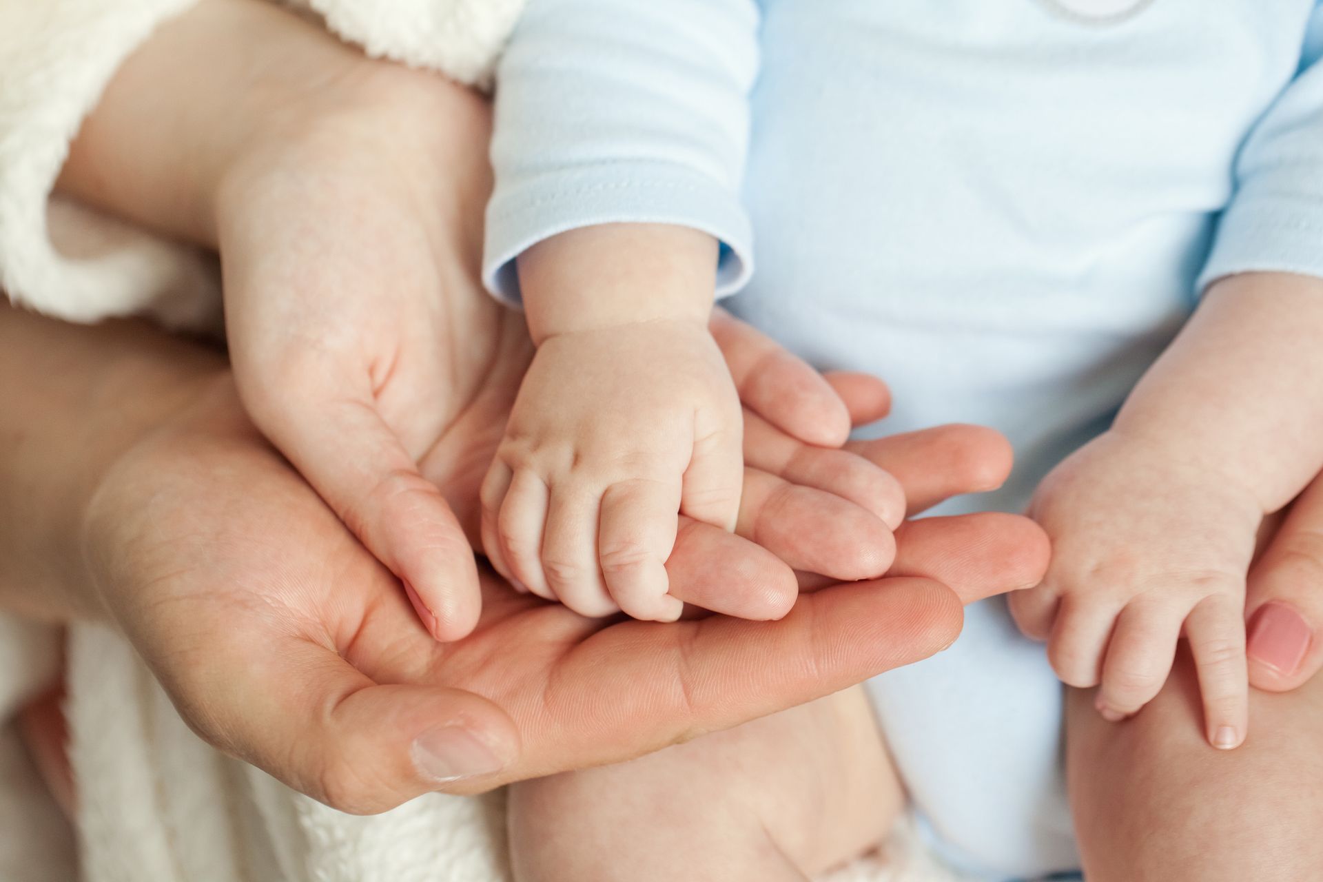 Baby's small hands resting on adult's hand, wearing a blue onesie.