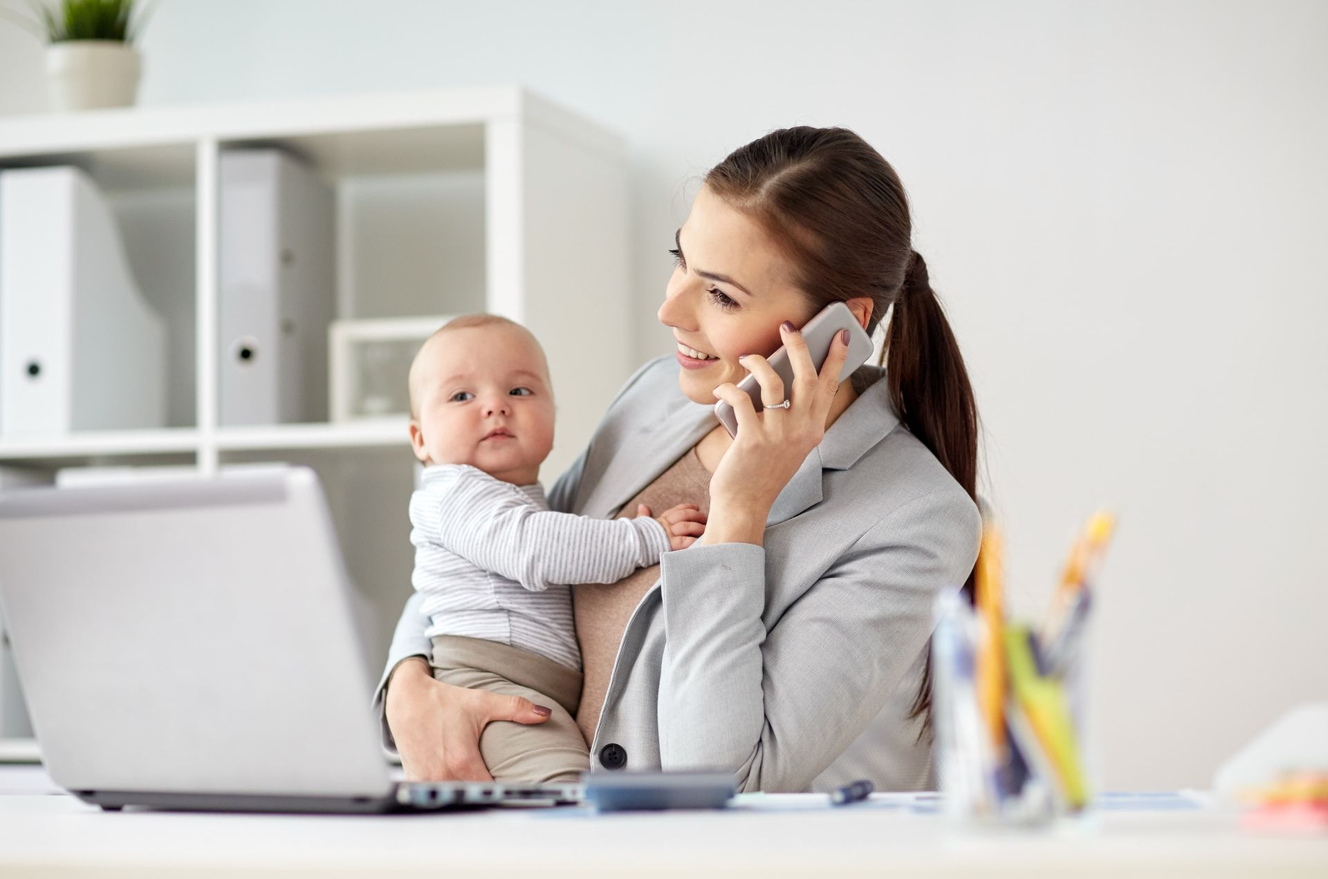 Woman in a blazer holds a baby, smiles, and talks on the phone while working on a laptop at a desk.