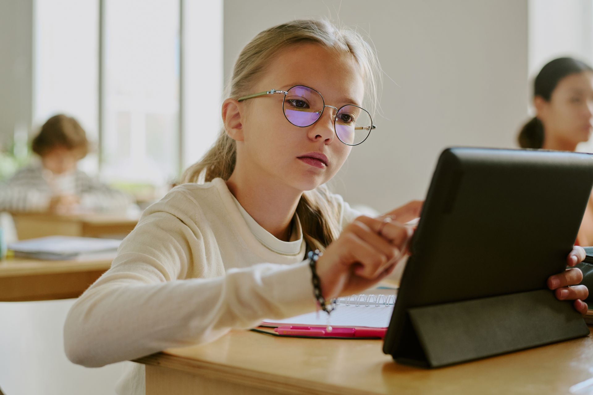 Student using a tablet at a desk in a classroom, with notebooks and classmates in the background. Student using a tablet at a desk in a classroom, with notebooks and classmates in the background.