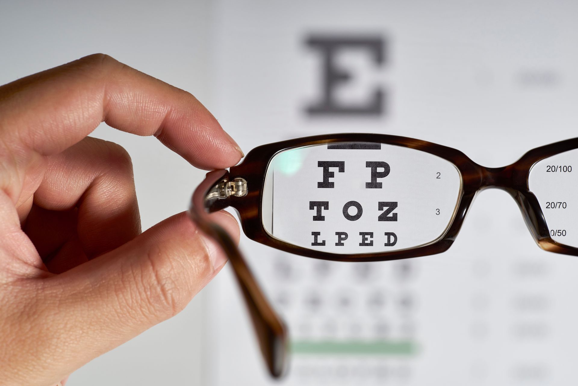 Hand holding eyeglasses in front of an eye chart, with the chart letters magnified through the lens Hand holding eyeglasses in front of an eye chart, with the chart letters magnified through the lens