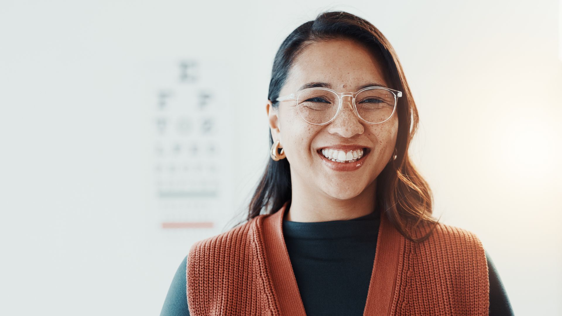 Smiling person in glasses and rust-colored sweater standing indoors against a bright background Smiling person in glasses and rust-colored sweater standing indoors against a bright background