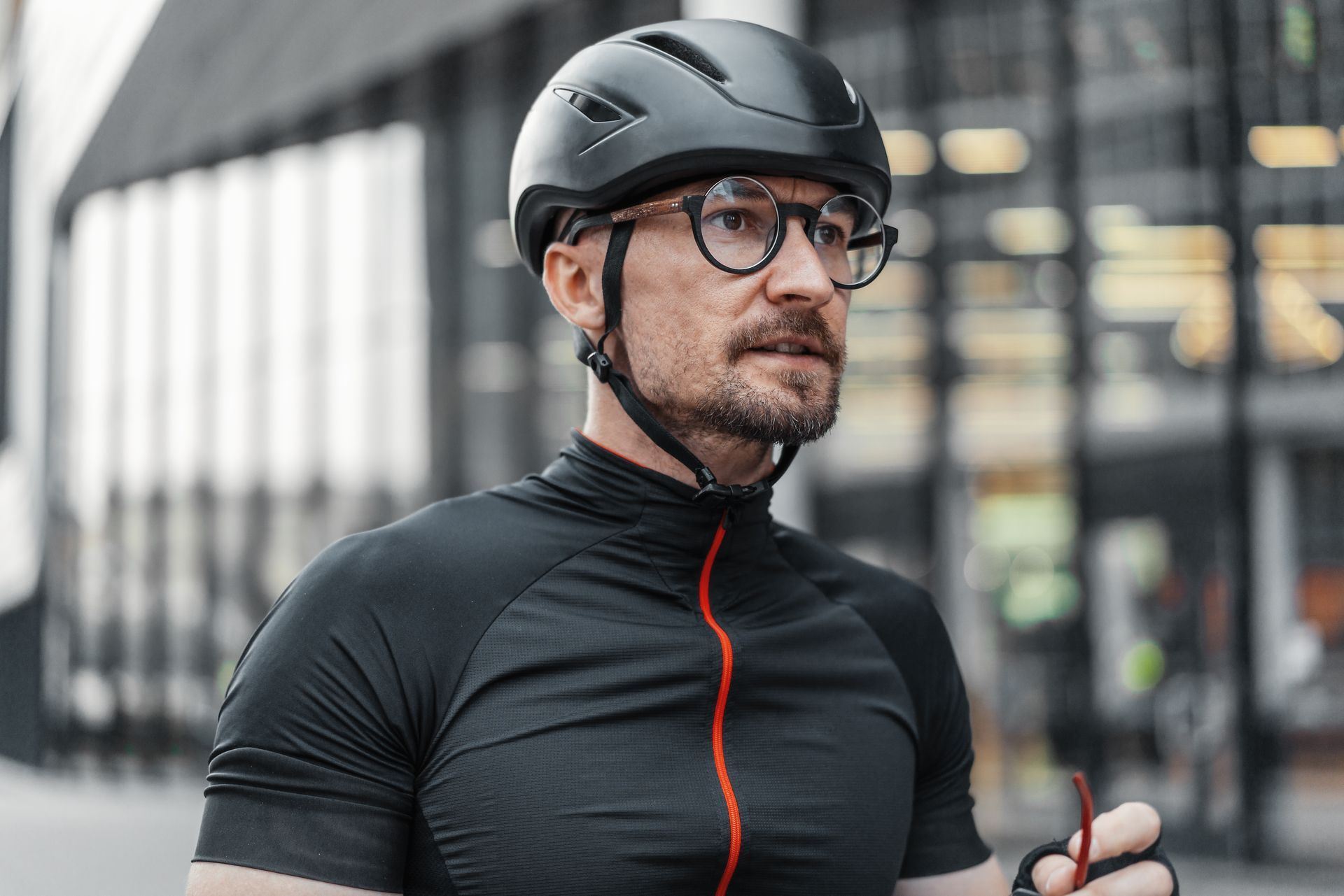 Cyclist in black helmet and jersey, standing outdoors in an urban setting with blurred buildings behind him Cyclist in black helmet and jersey, standing outdoors in an urban setting with blurred buildings behind him
