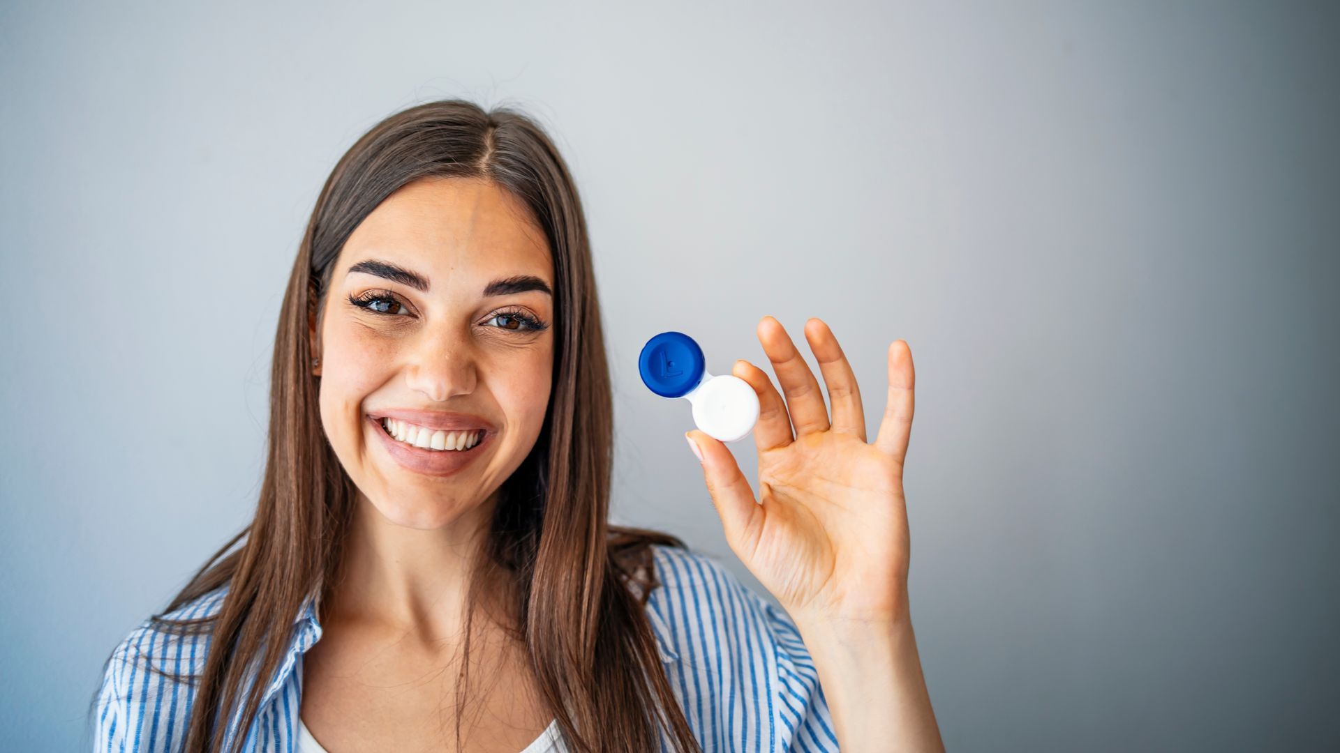 Smiling person holding a blue and white contact lens case against a gray background Smiling person holding a blue and white contact lens case against a gray background