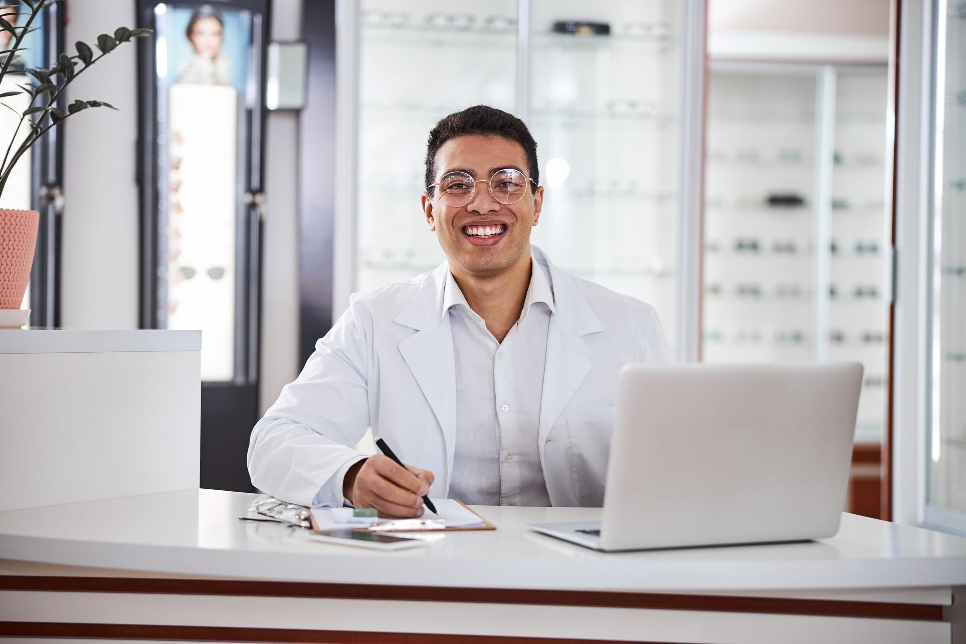 Person in a white coat smiling at a desk with a laptop and clipboard in a bright office