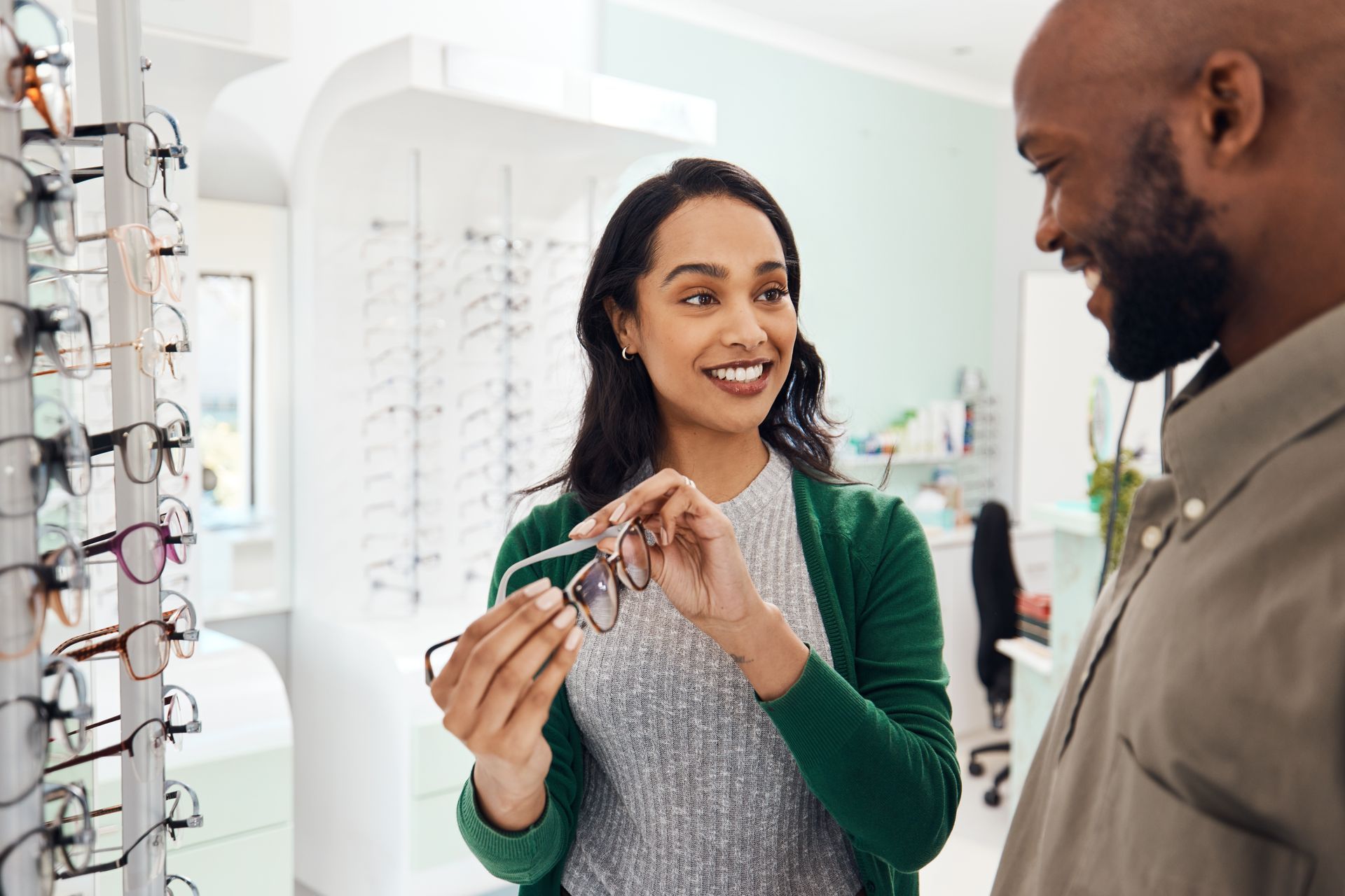 Two people discussing eyeglass frames in a bright optical store, with one holding a pair up to show the other. Two people discussing eyeglass frames in a bright optical store, with one holding a pair up to show the other.