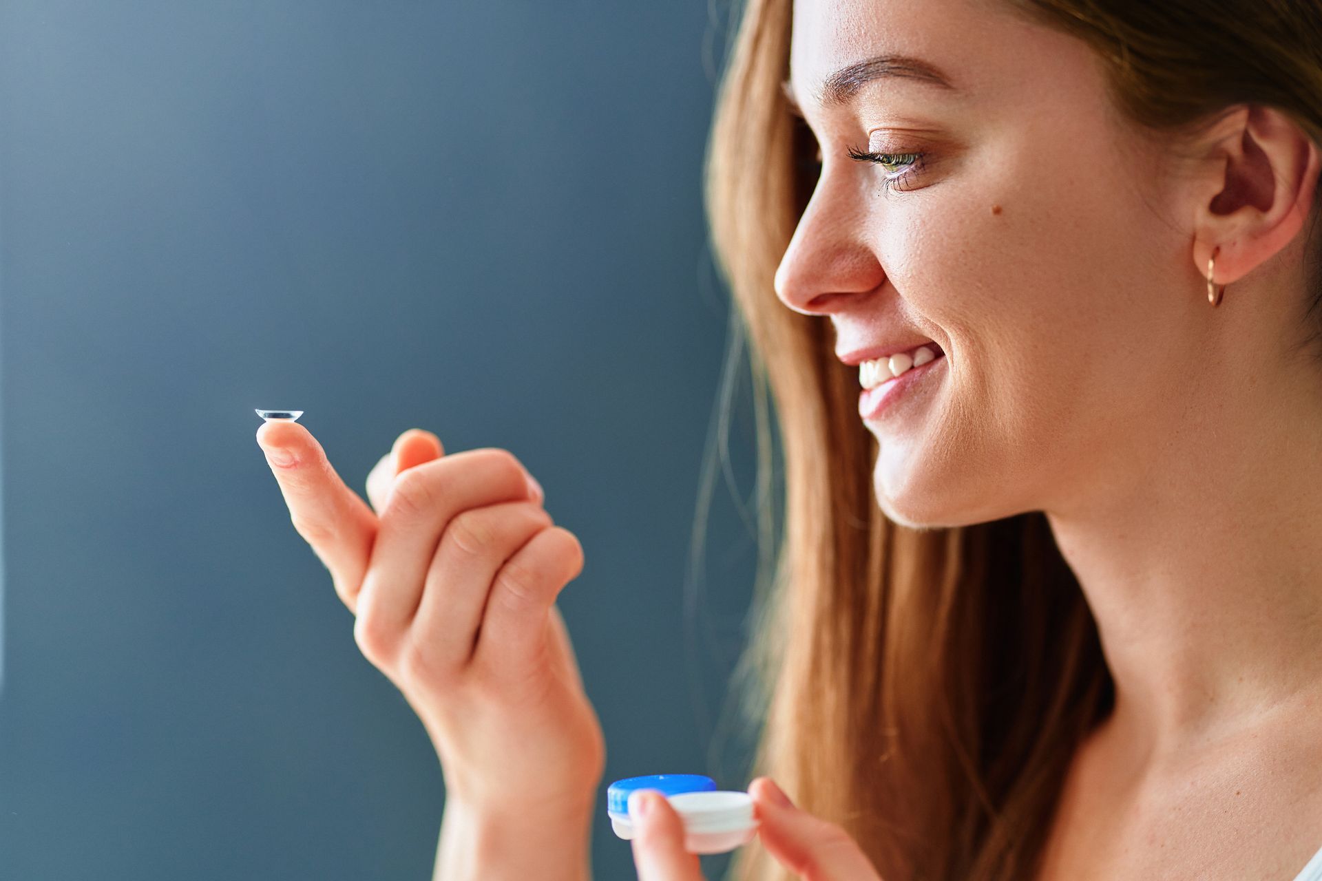 Woman smiling while holding a contact lens and lens case against a blue background Woman smiling while holding a contact lens and lens case against a blue background