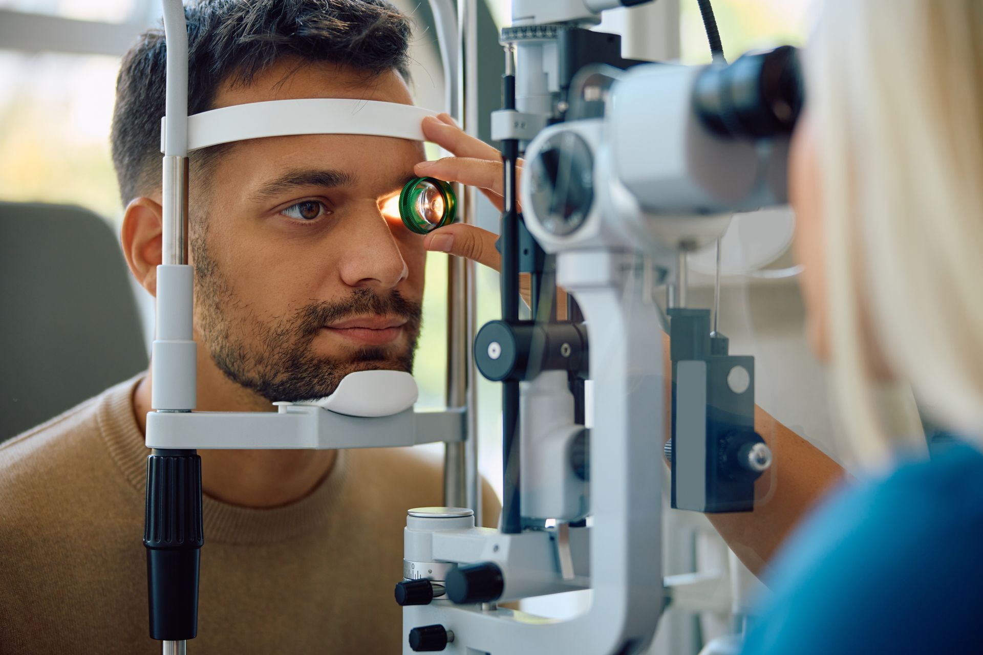 Patient undergoing an eye exam at a slit lamp while a clinician adjusts the equipment Patient undergoing an eye exam at a slit lamp while a clinician adjusts the equipment