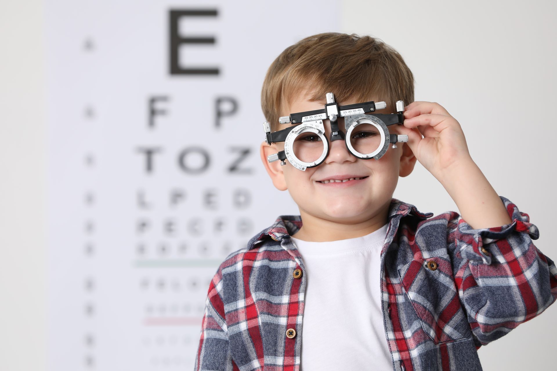 Child holding an eye chart testing device in a vision exam room, with a chart in the background