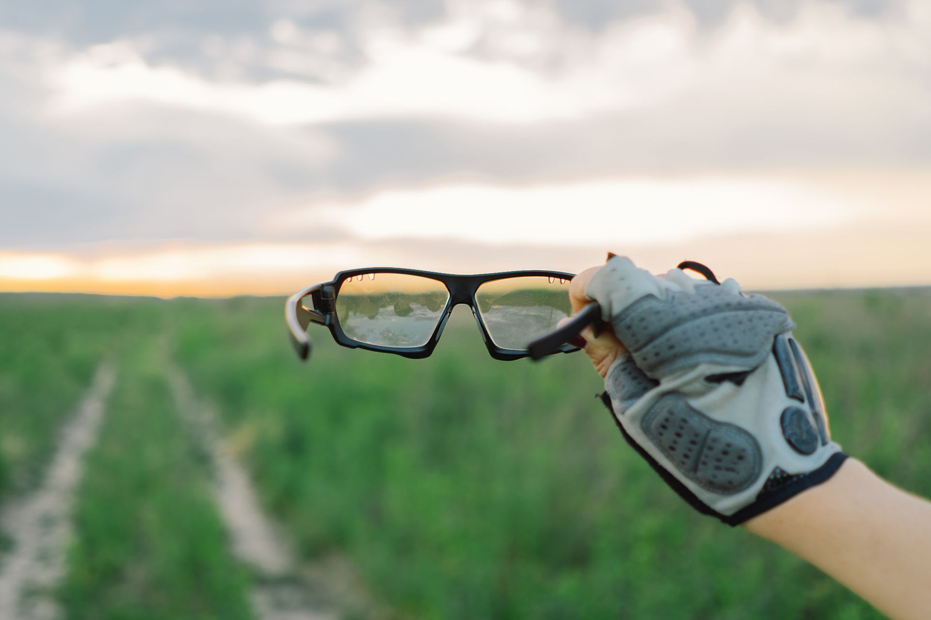 Gloved hand holding black eyeglasses over a green field at sunset