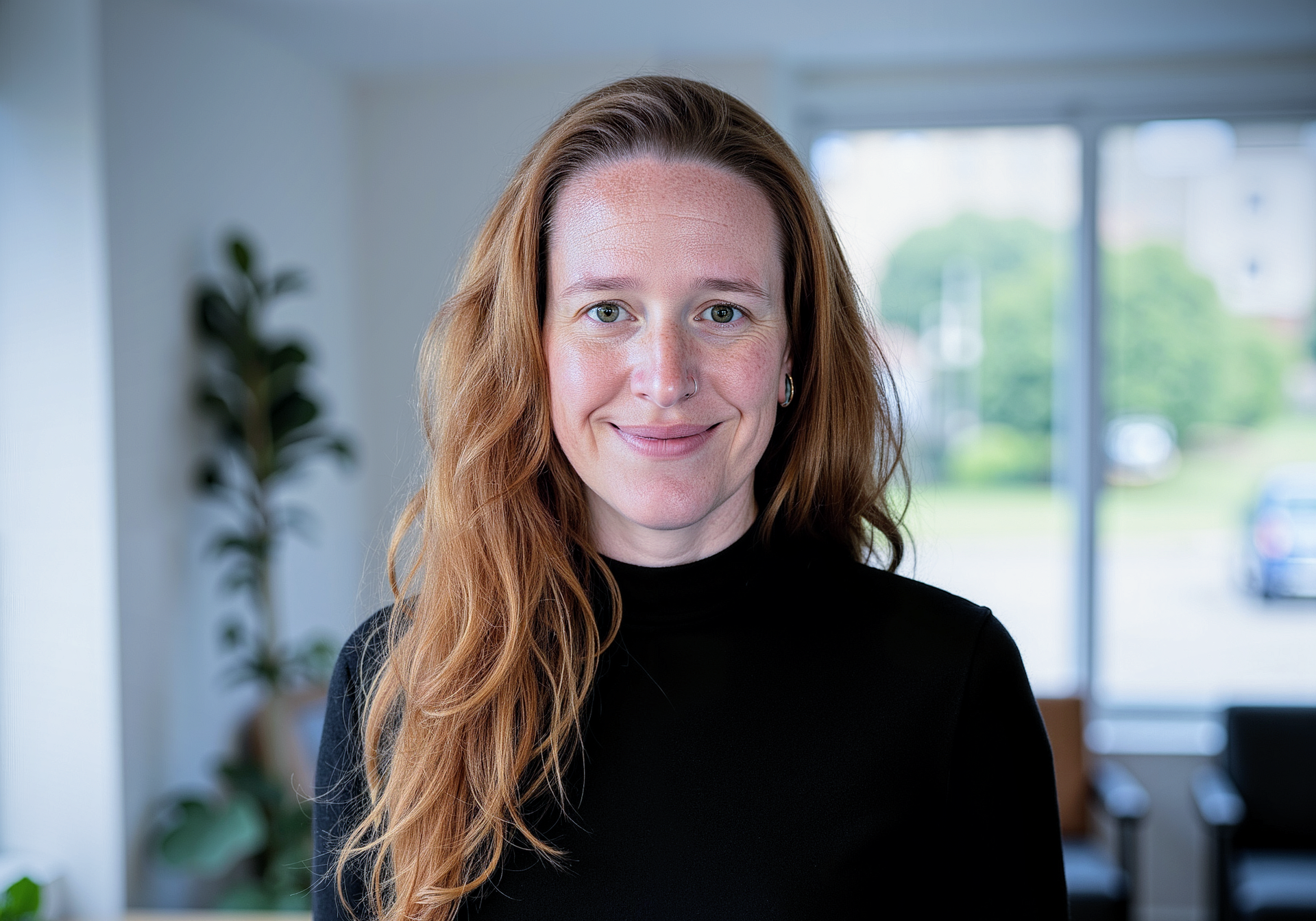 Woman with long red hair smiles, wearing a black turtleneck, in a bright room with a window.