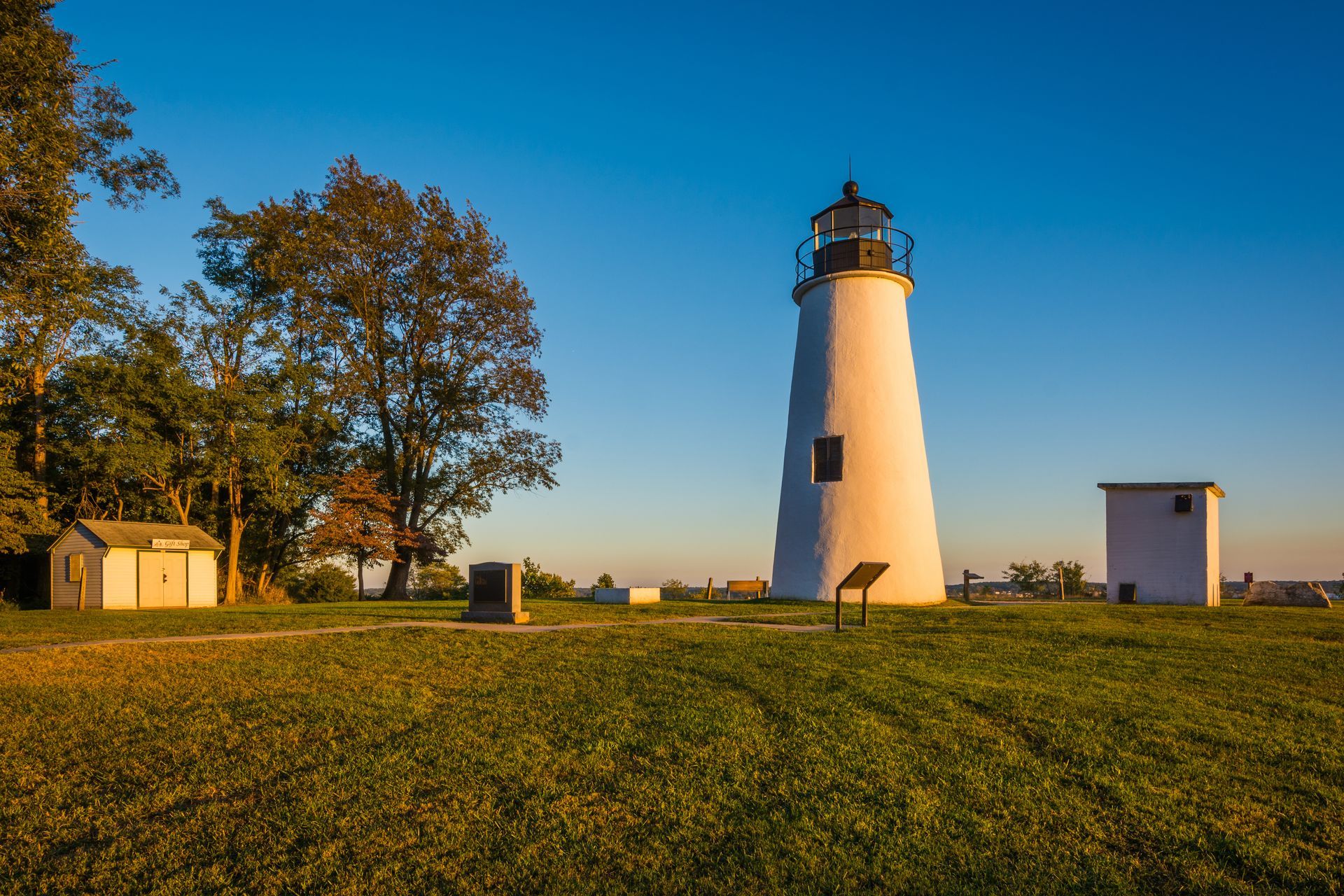 Lighthouse on a grassy hill at sunset, with trees and small buildings under a clear blue sky