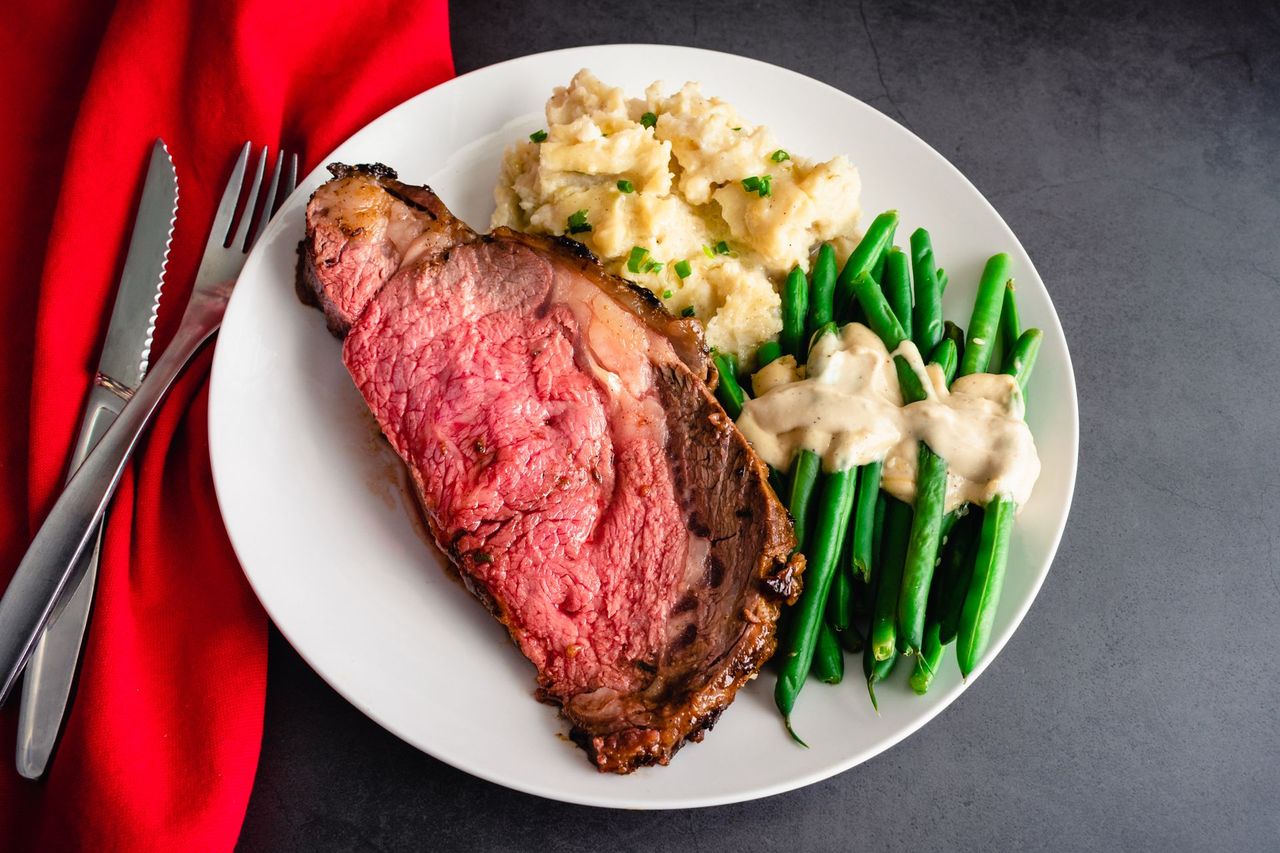 Plate with sliced roast beef, mashed potatoes, and green beans on a white dish over a red tablecloth