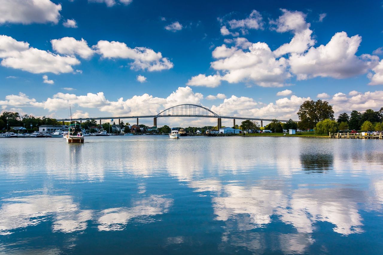 Wide river with bridge, blue sky, scattered clouds, and reflections on calm water