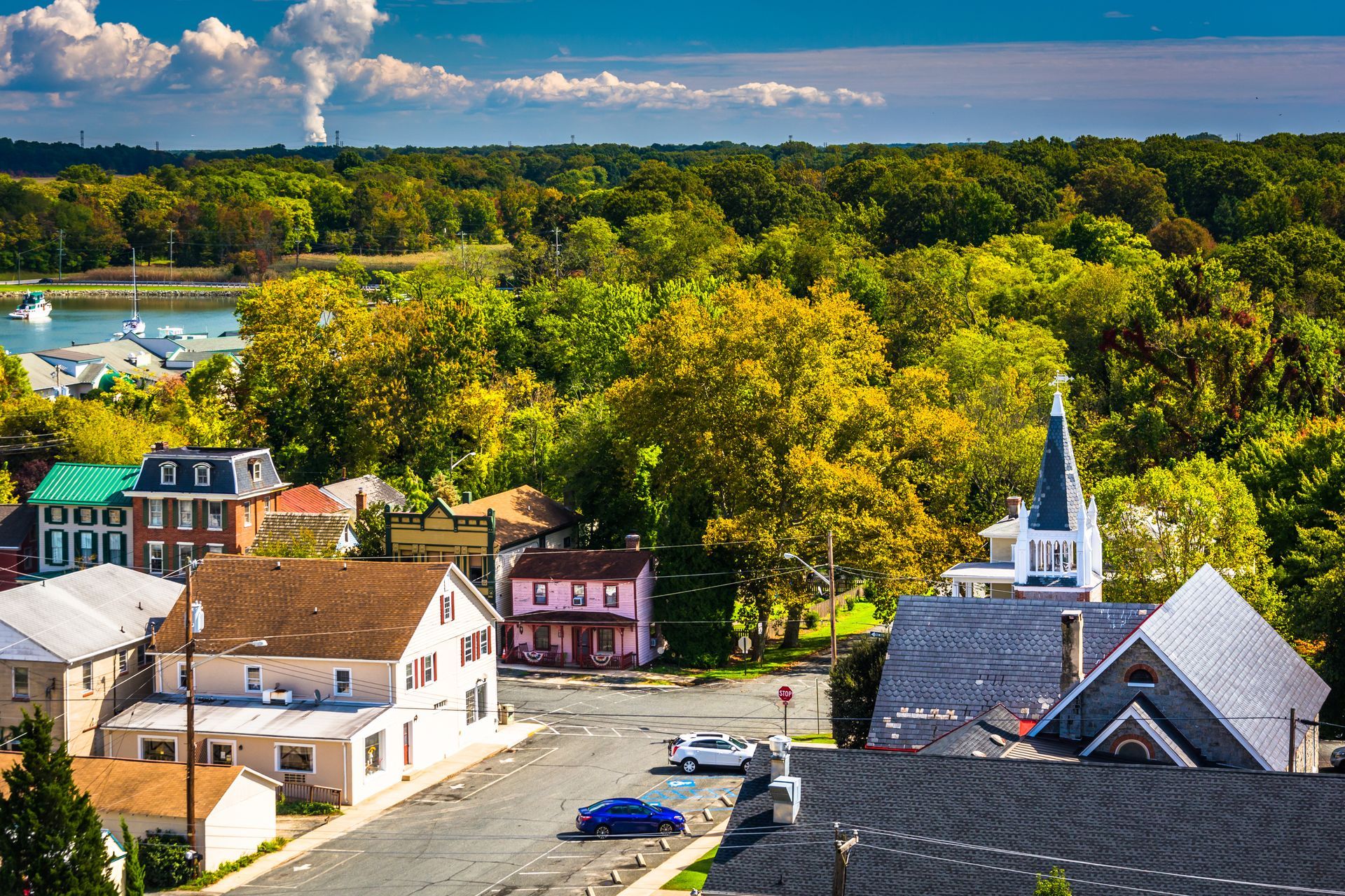Colorful village rooftops beside a road, with a white church steeple and lush green forest beyond.