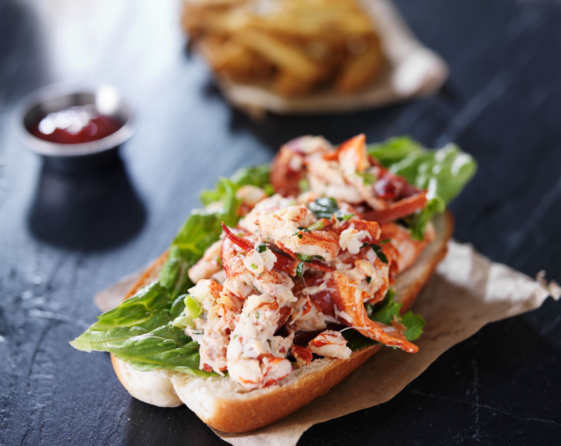Lobster roll on a bun with lettuce and red garnish, served on dark table with fries and dipping sauce.