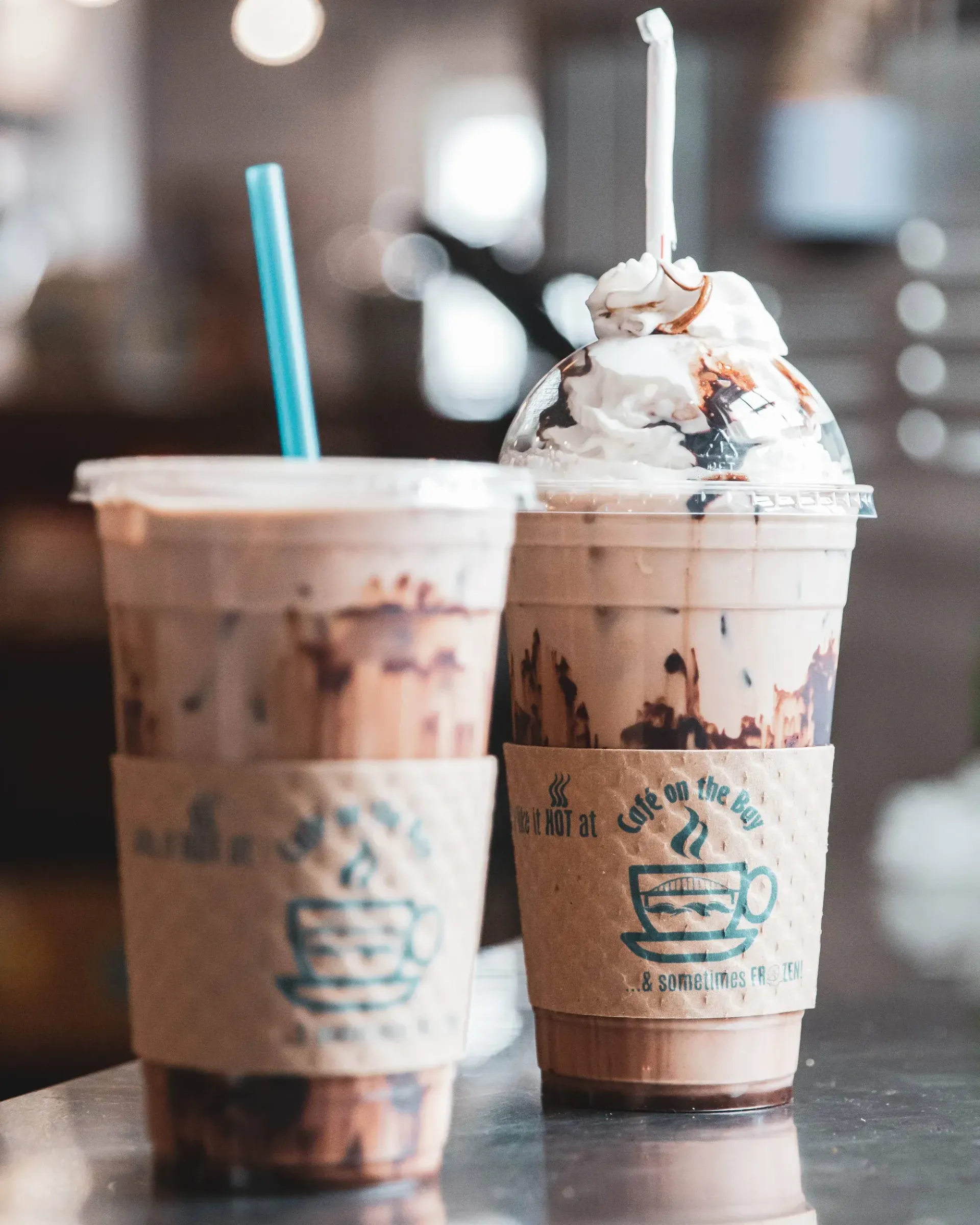 Two iced coffee drinks on a table, one topped with whipped cream and chocolate drizzle
