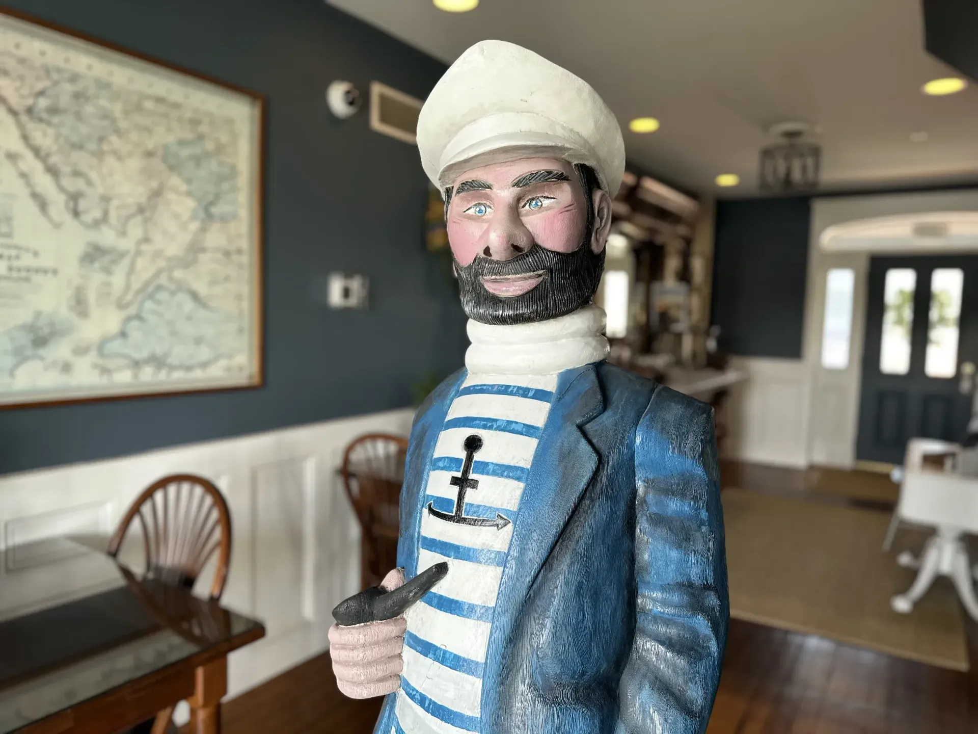 Bearded figurine in a blue coat and white hat standing in a hallway with framed art and chairs