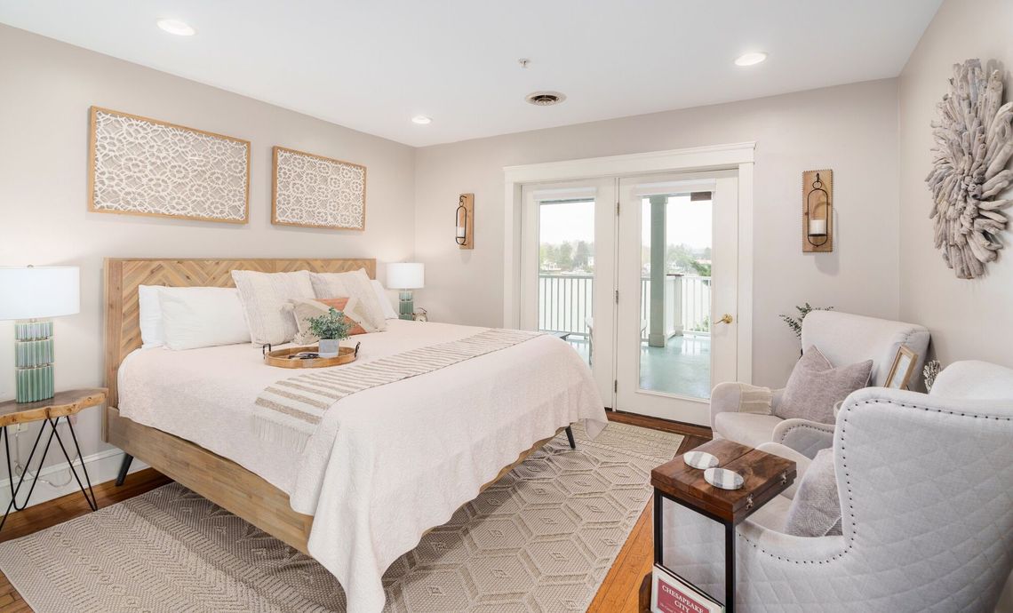 Bright bedroom with a white bed, gray chair, woven rug, and French doors leading outside