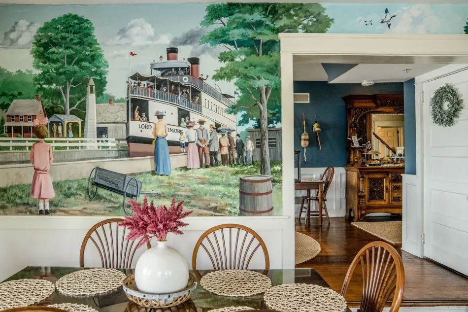 Dining room with wicker chairs and floral decor beside a mural of a waterfront street scene.