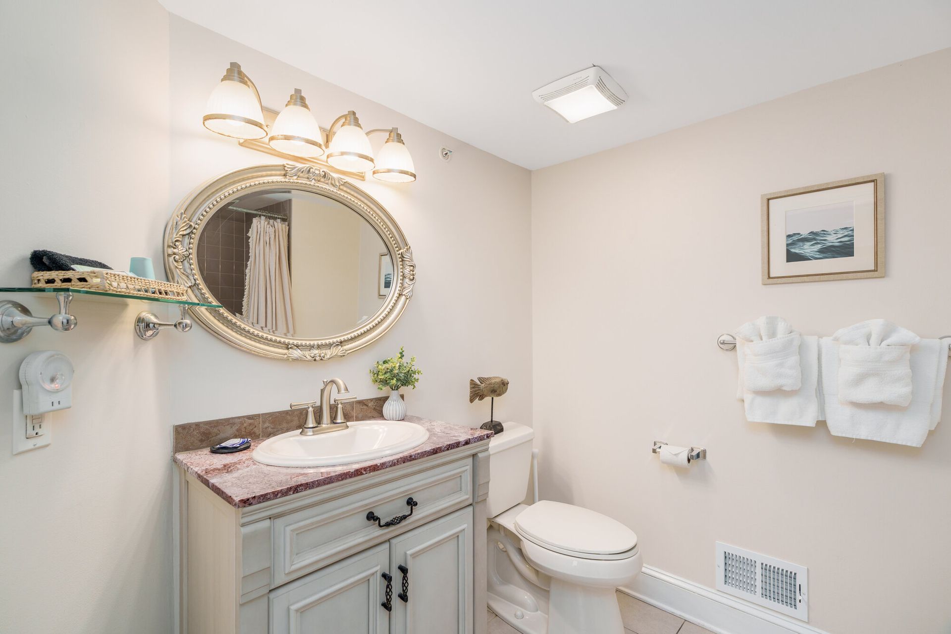 Bright bathroom with ornate mirror, vanity, toilet, wall towels, and framed art.