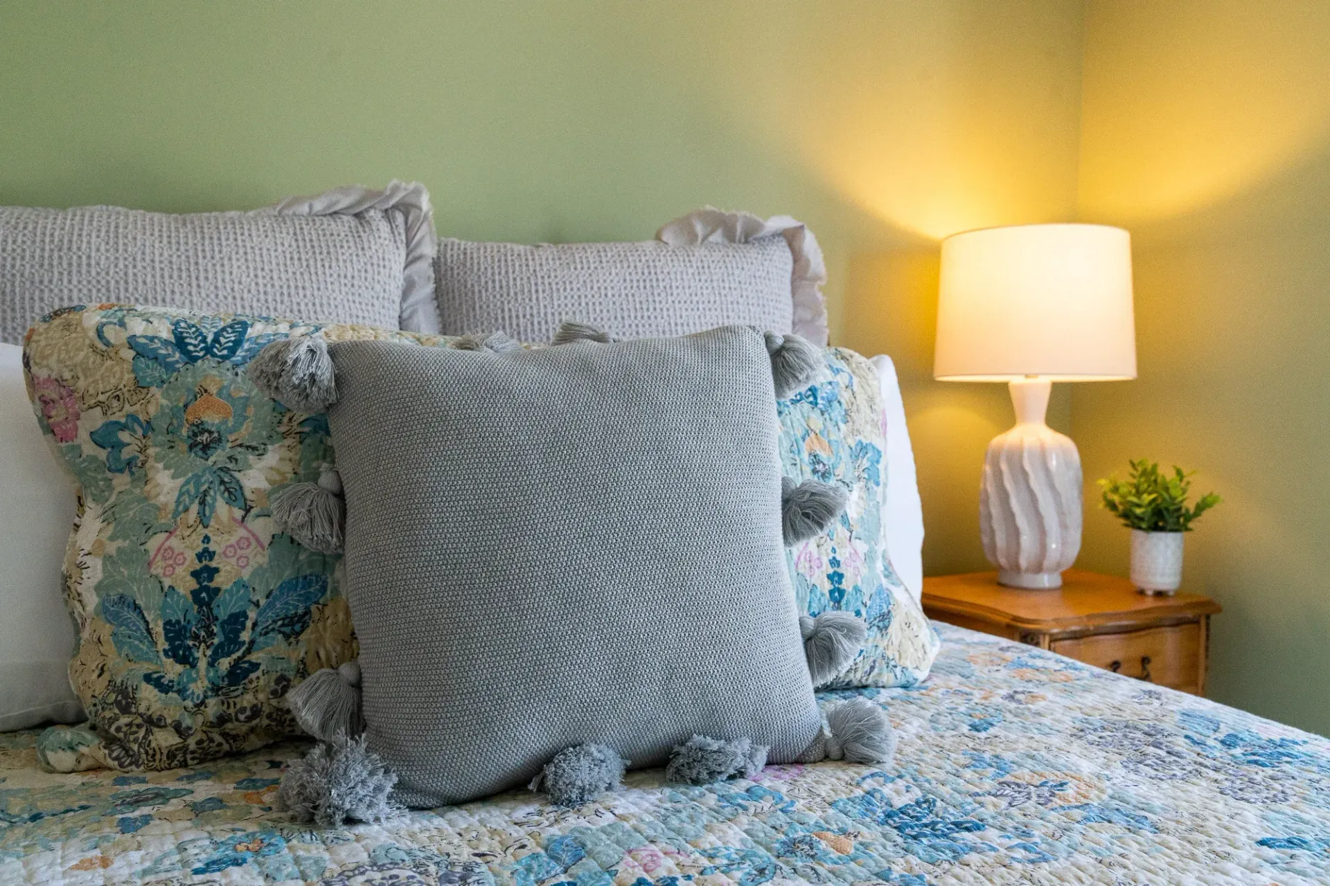 Floral bed with gray pillows beside a lit lamp on a wooden nightstand in a cozy bedroom