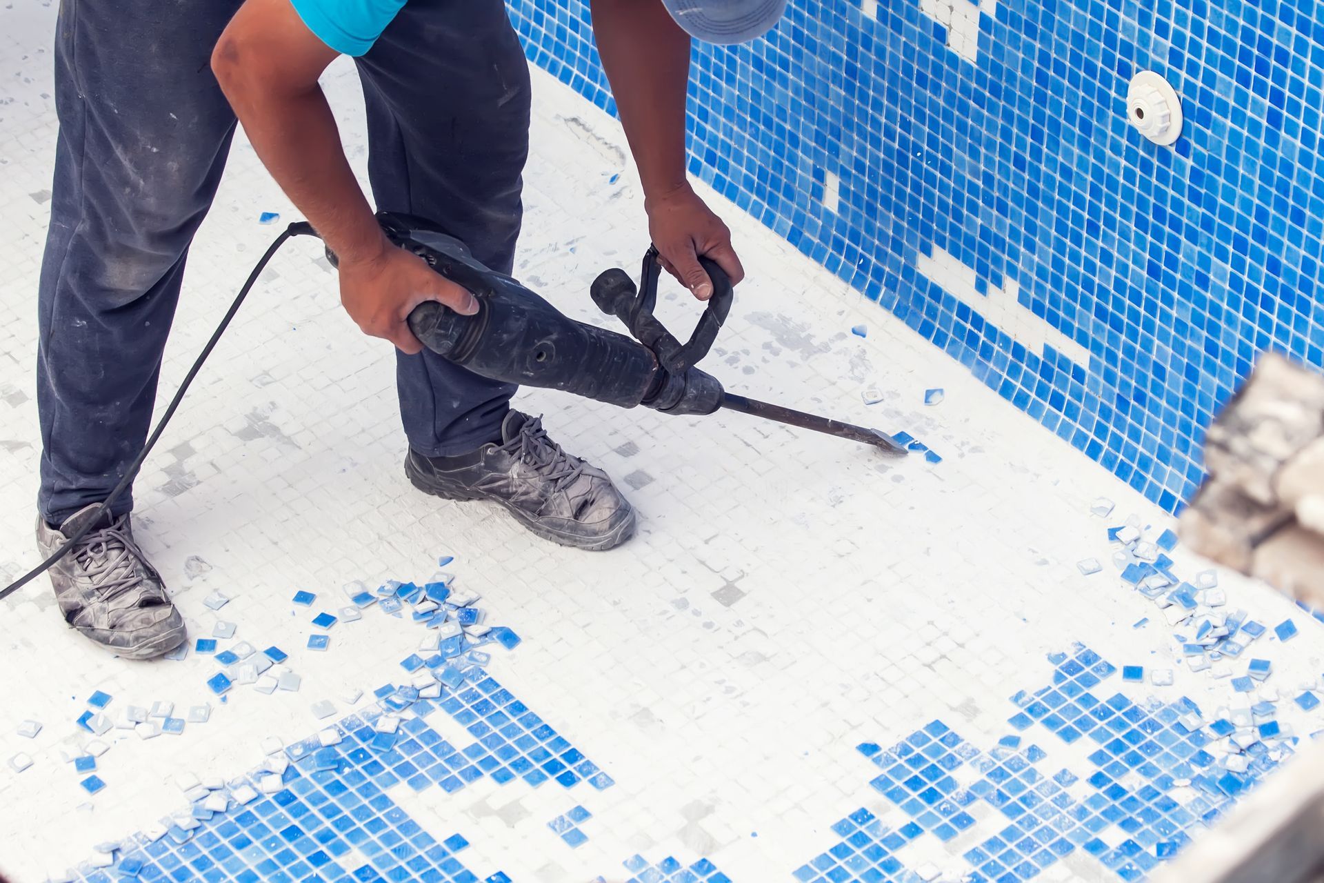 A worker using a power tool to remove blue and white mosaic tiles from a pool surface. A worker using a power tool to remove blue and white mosaic tiles from a pool surface.