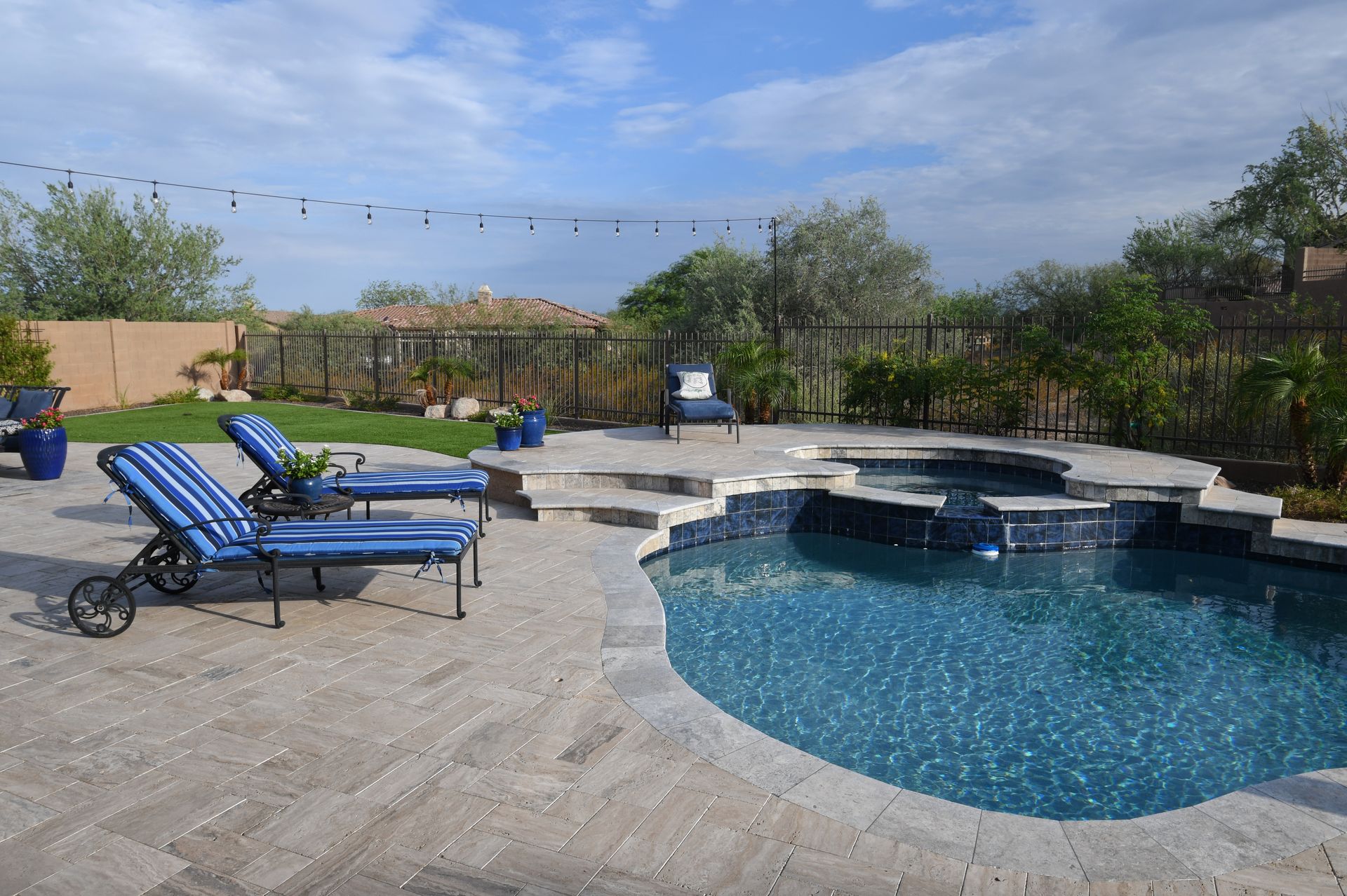 Backyard pool with blue lounge chairs, stone patio, and trees under a clear sky Backyard pool with blue lounge chairs, stone patio, and trees under a clear sky
