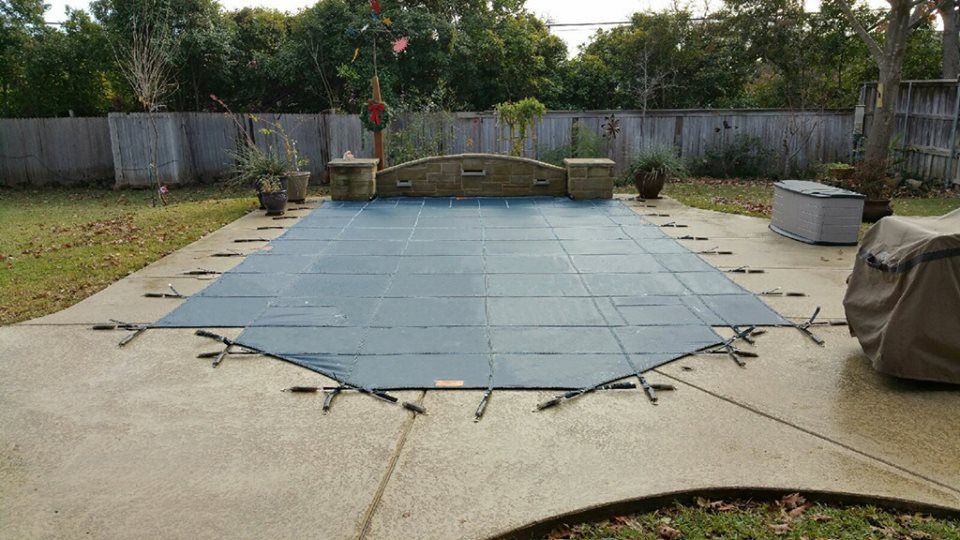 Pool covered with a dark blue safety cover, surrounded by a concrete patio and a wooden fence.