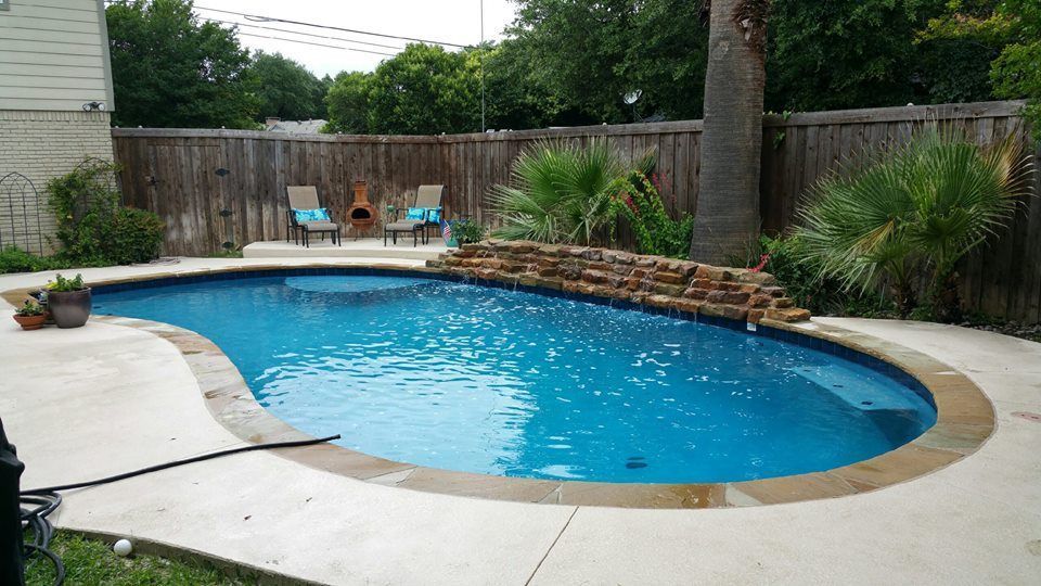 A backyard pool with a waterfall feature surrounded by concrete and a wooden fence.